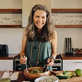 Woman in green shirt in the kitchen smiling and tossing a salad
